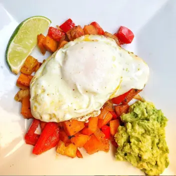 plate of sweet potato hash topped with over easy egg and a side of mashed avocado and lime wedge