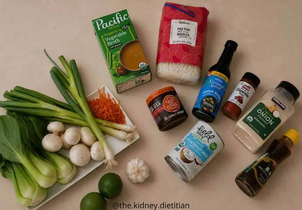 More low sodium ramen ingredients on counter: low sodium vegetable broth, rice noodles, chili crisp, vegetables, coconut milk and spices