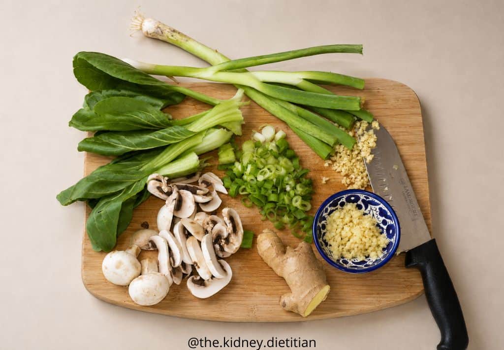 Cutting board with low sodium ramen ingredients on it - green onions, baby bok choy, mushrooms, ginger and garlic